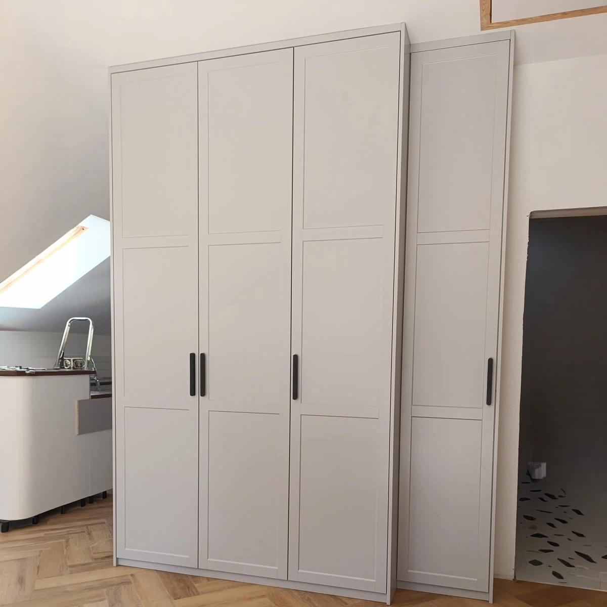 Einbauschrank nach Maß mit hellgrauen Rahmenfronten und schwarzen Stangengriffen, dreitürig mit schmalem Zusatzelement, in einem Schlafzimmer mit Dachschräge und Fischgrätparkett.